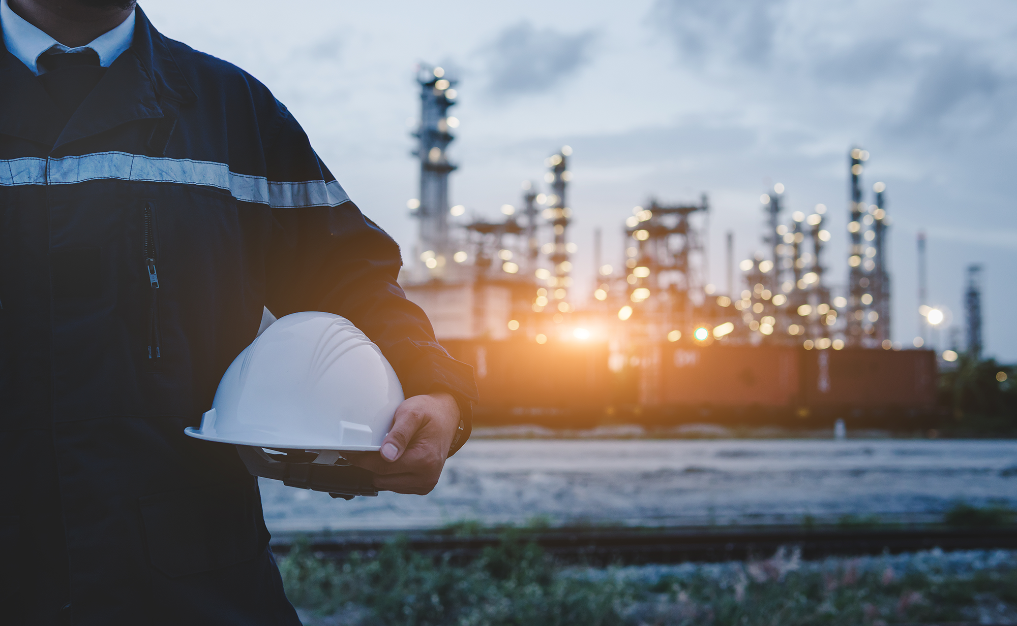 person standing in front of a refinary with a hardhat in this hand